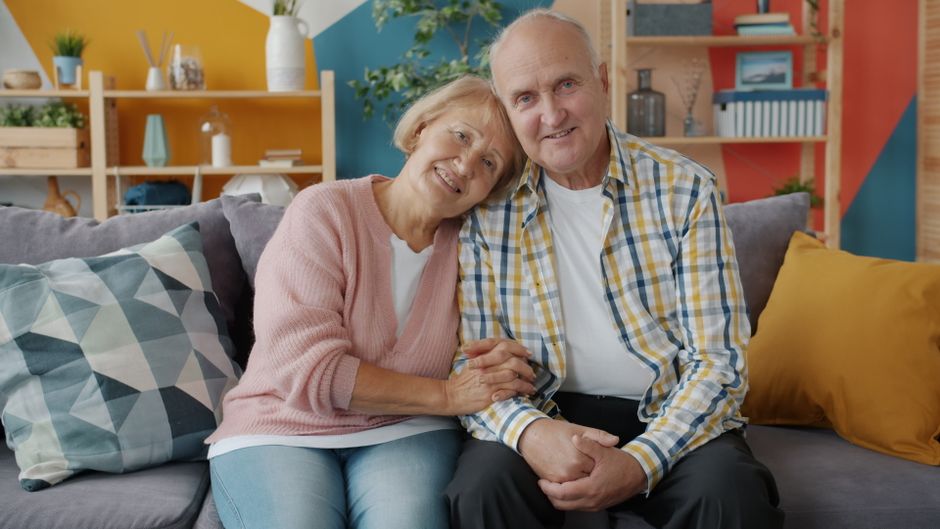 Un couple de personnes âgées souriantes, assises côte à côte sur un canapé confortable dans un salon coloré.